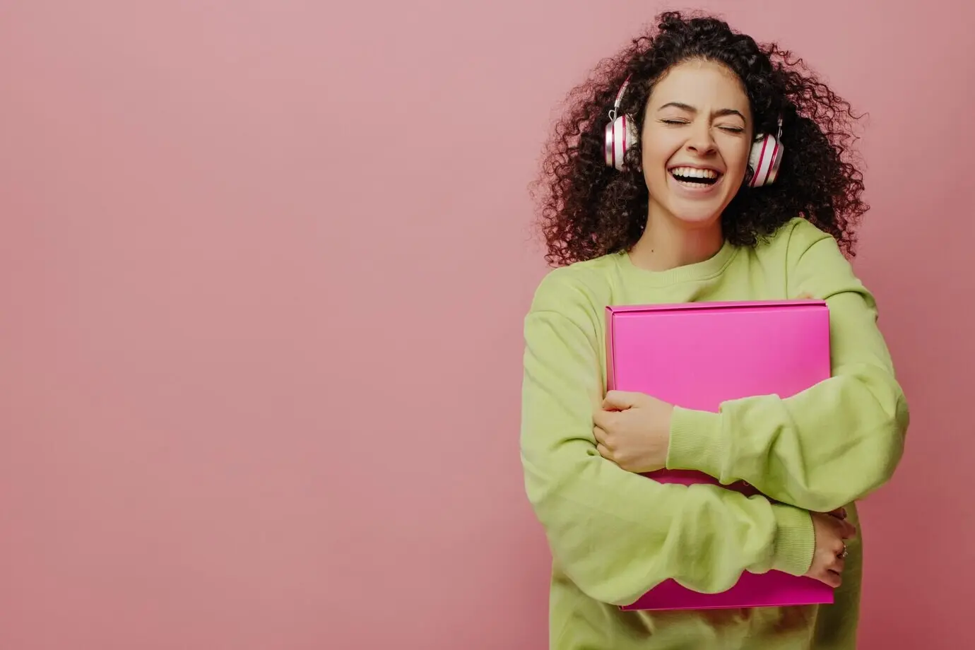 Ein junges, hellhäutiges, lockiges brünettes Mädchen mit Kopfhörern lacht mit geschlossenen Augen, hält eine Schachtel fest an die Brust und hört ihre Lieblingsmusik vor einem Hintergrund mit Platz für Text – Konzept des Genießens des Augenblicks.