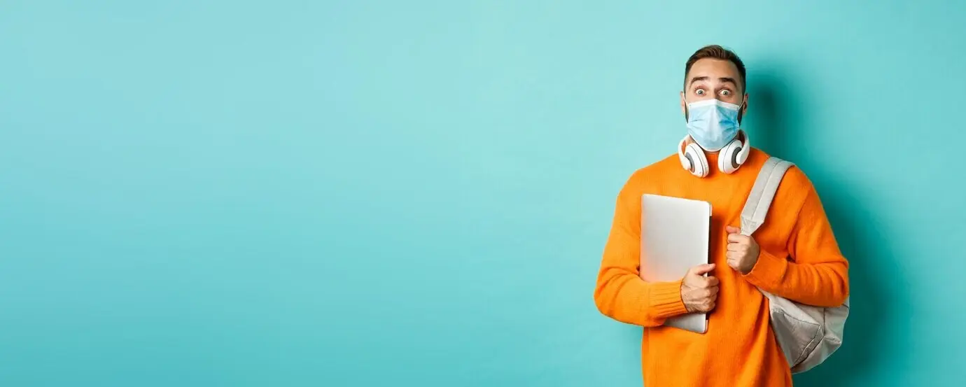 Attraktiver kaukasischer Mann mit Kopfhörern und Rucksack, der einen Laptop hält, eine medizinische Maske trägt und blickt.